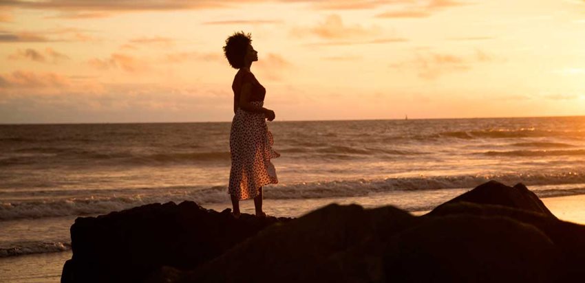 Mujer parada en una roca y en el fondo la playa y el mar, al atardecer.
